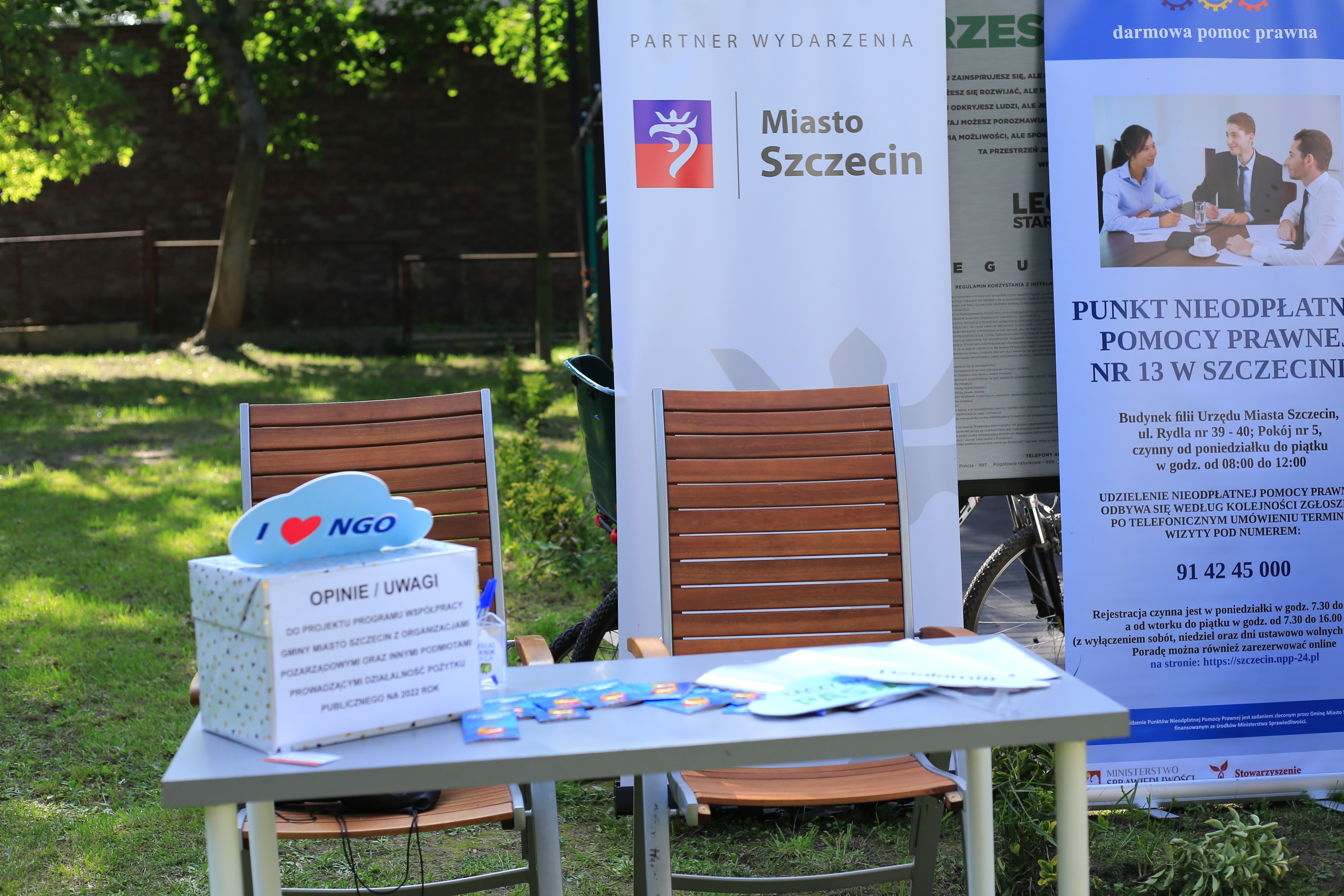 Stolik w parku, na którym znajduje się urna z napisem "opinie, uwagi". Na urnie znaczek i love NGO. W tle roll-upy Miasta szczecin oraz Punktu Nieodpłatnej Pomocy Prawnej.