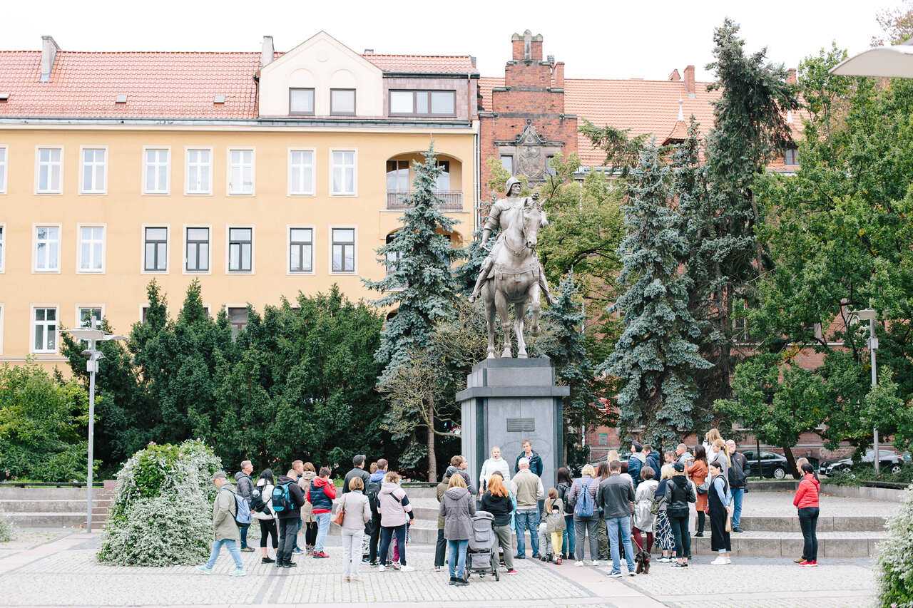 Zdjęcie organizatora, przedstawiające grupę zwiedzających, stojących na Placu Lotników pod pomnikiem Bartolomea Colleoniego. W tle kamienice w centrum Szczecina.